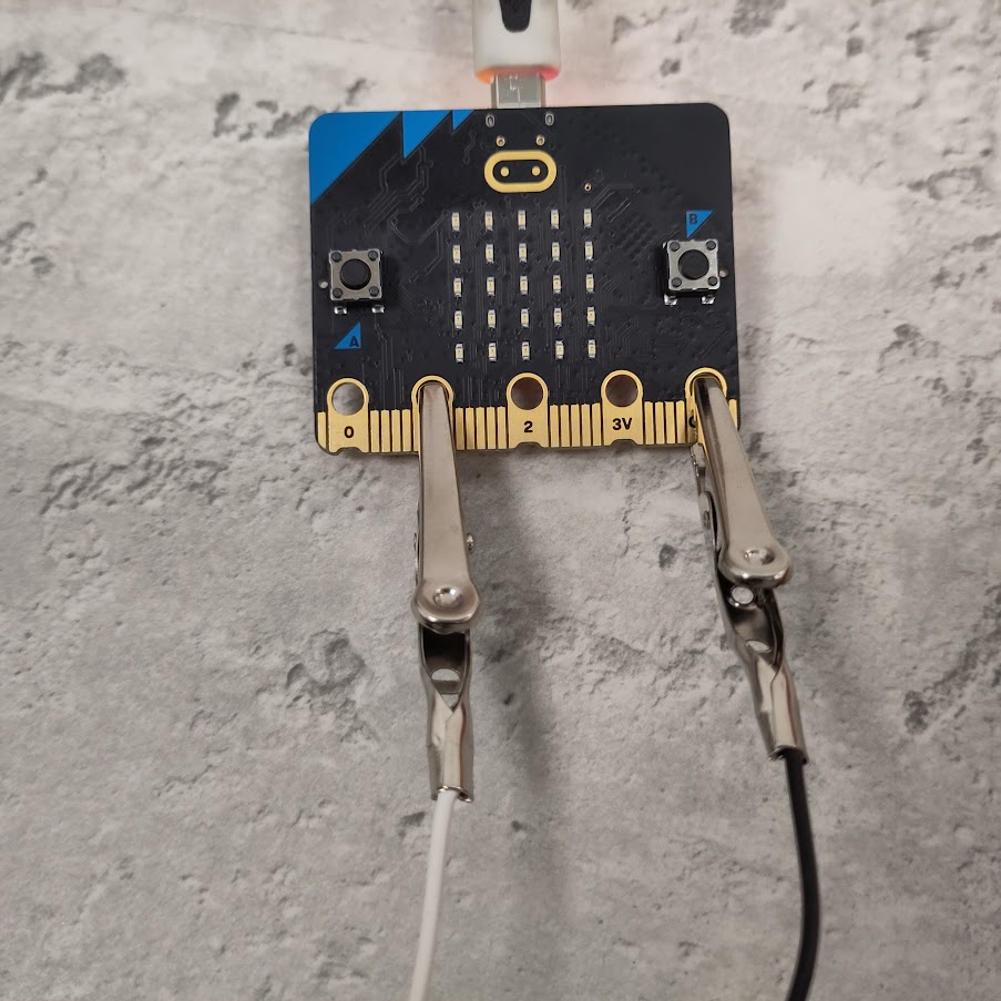 photograph of a BBC micro:bit. A crocodile clip with a white wire is attached to the gold pin labelled 1, second from the left. A crocodile clip with a black wire is attached to gold pin labelled GND, furthest right.